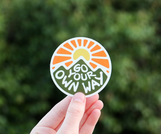 Squatchy Go Your Own Way vinyl sticker featuring a mountain and sun graphic held against a blurred green background
