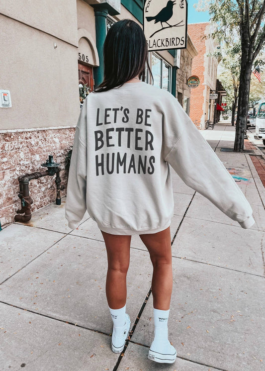 Person wearing a sweatshirt with 'Let's Be Better Humans' text on a sidewalk.