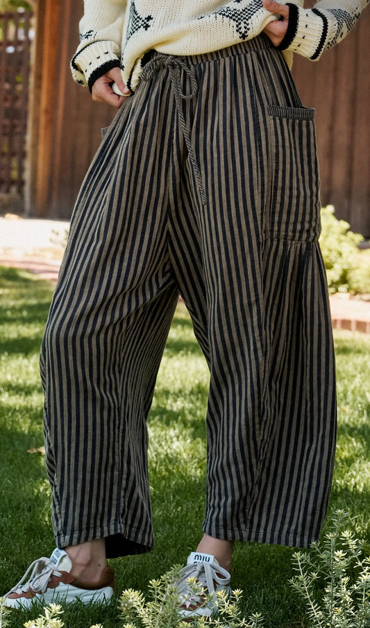 A person outdoors wearing loose, wide-leg striped pants in dark gray and beige, with a cream embroidered top and white sneakers, standing on grass in front of a wooden fence.