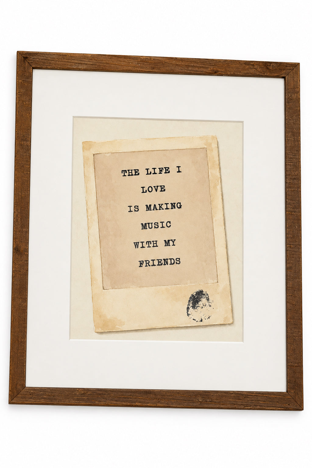 A square typographic art print featuring the text 'THE LIFE I LOVE IS MAKING MUSIC WITH MY FRIENDS' in black on a beige background, mounted on a larger white mat inside a clear plastic sleeve. The print is photographed on a wooden surface with a hand visible at the bottom edge.