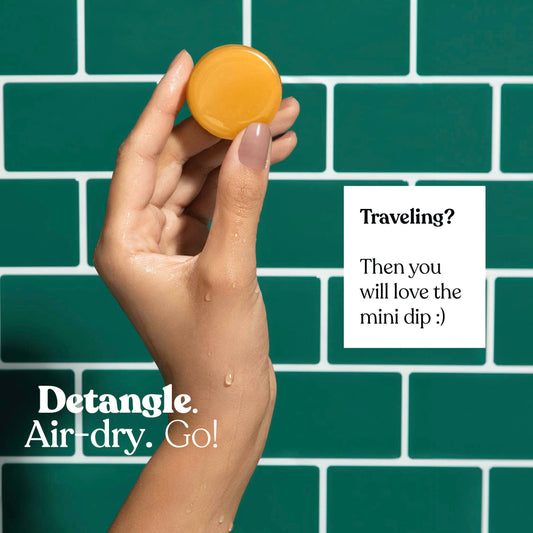 Hand holding a round orange Dip Mini Conditioner and After Swim Detangler bar against a green tiled background