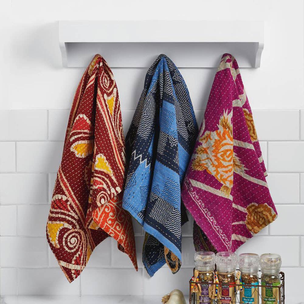Three SERRV International Kantha dish towels in red blue and pink hanging on a white wall rack