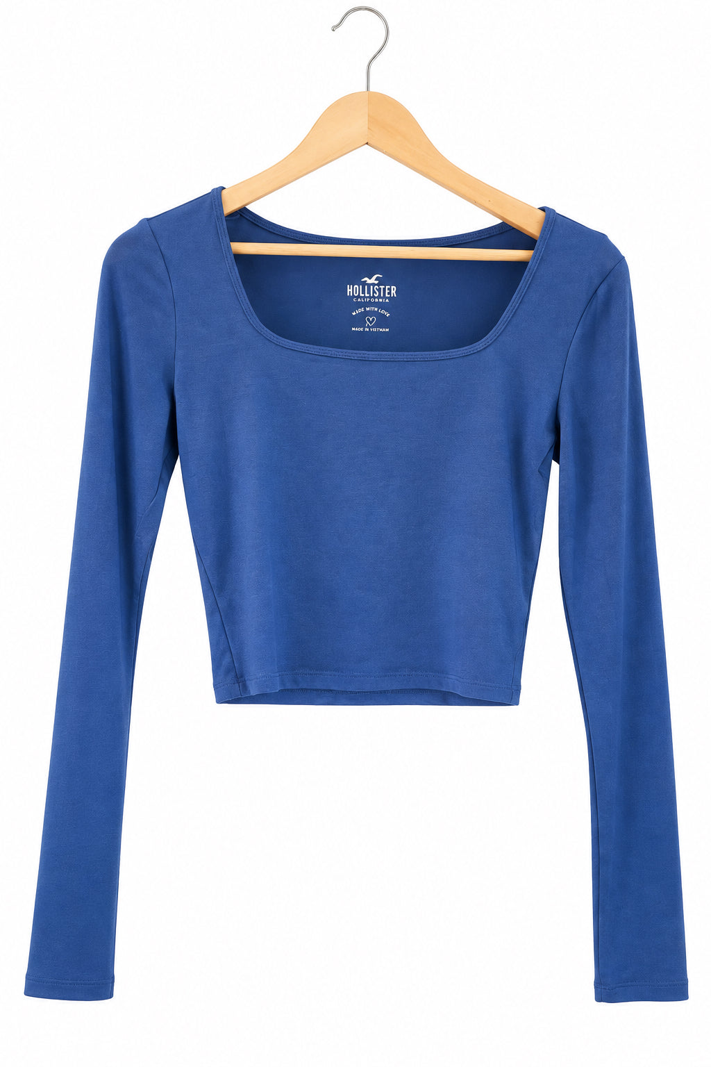 A royal blue long-sleeve scoop-neck crop top hanging on a wooden hanger in front of a dark gray paneled door.