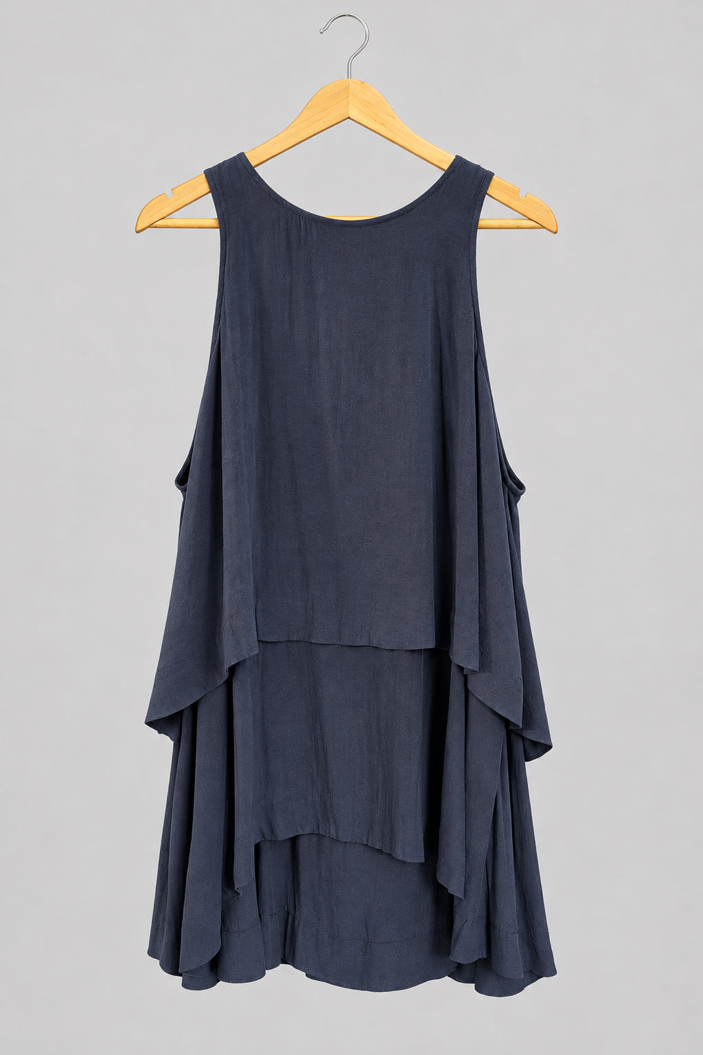 A sleeveless, tiered high-low tank top in a muted slate gray-blue color, hanging on a wooden hanger against a dark gray paneled door.