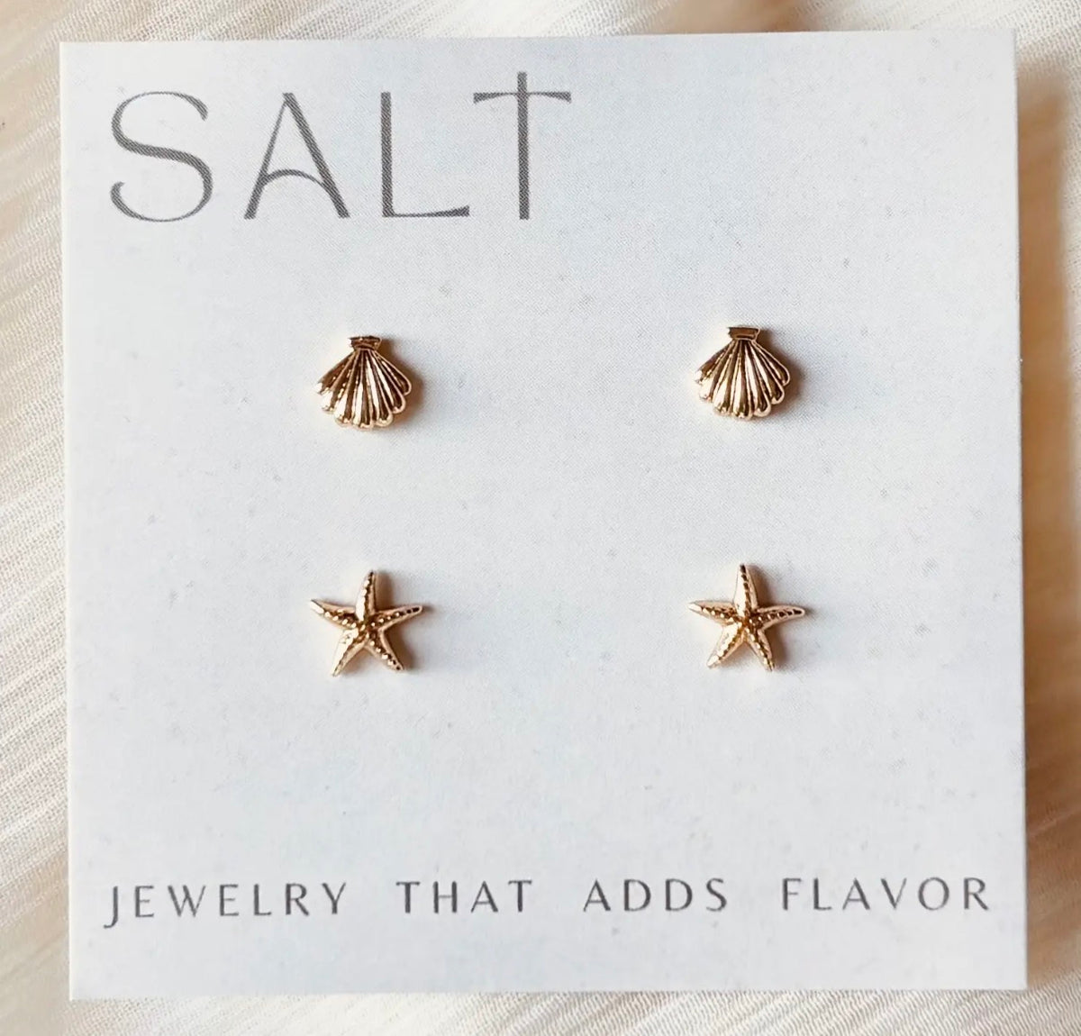 Two pairs of gold-tone coastal stud earrings featuring textured seashells and starfish, displayed on a white jewelry card.