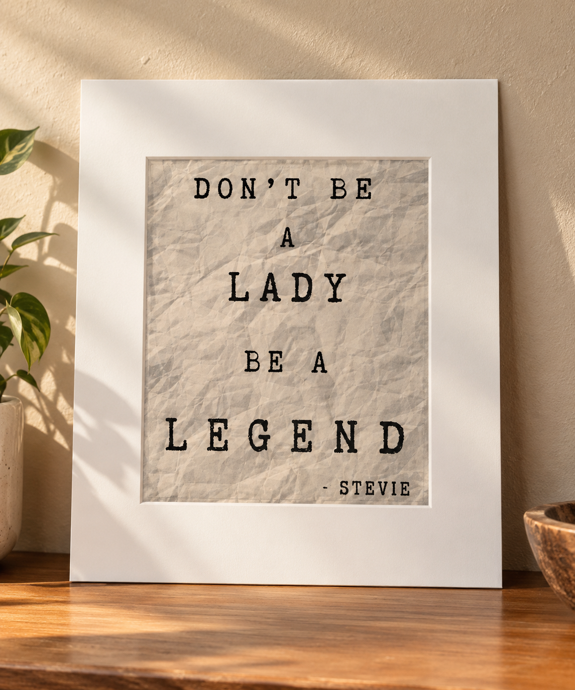 A framed typographic wall art print leaning on a wooden surface against a beige wall, with a plant and bowl partially visible. The print reads “DON’T BE A LADY BE A LEGEND - STEVIE” on a textured light beige background with a wide white frame.
