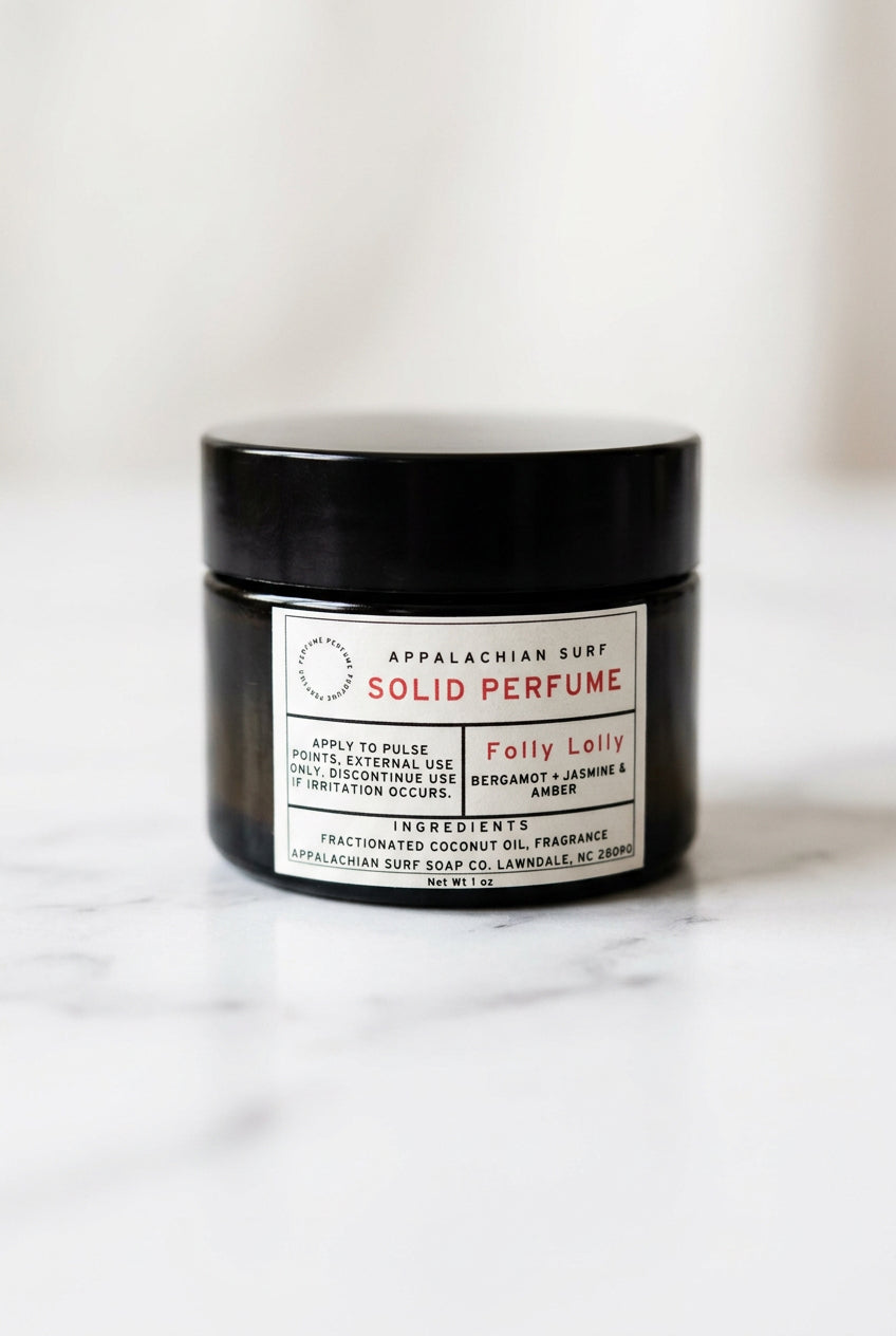A jar of Appalachian Surf Solid Perfume labeled 'Folly Lolly' sits on a marble countertop with a blurred wooden shelf and soap bars in the background.
