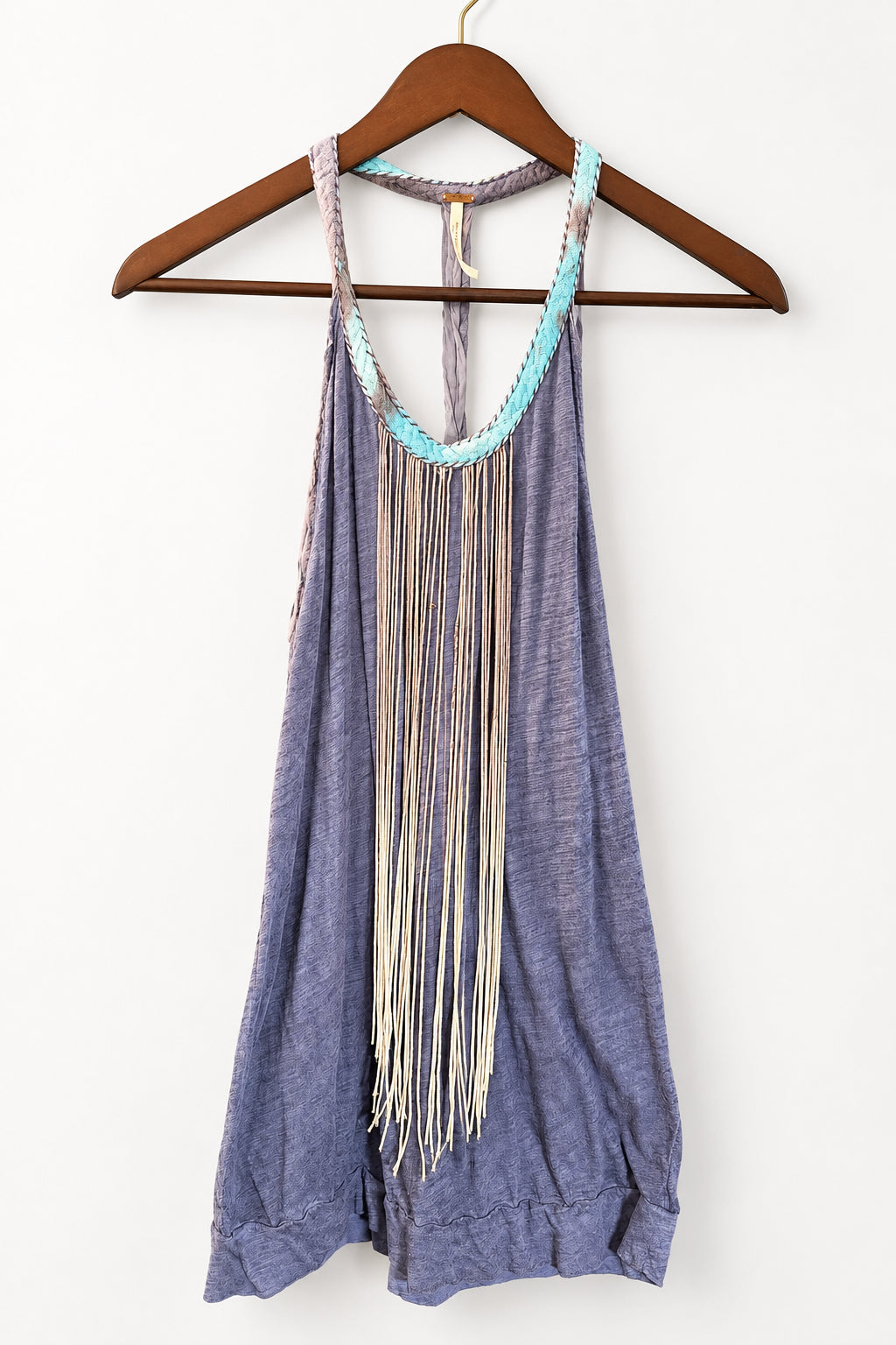 A sleeveless dusty purple fringed dress hanging on a wooden hanger against a brown paneled door. The dress features a light blue and white beaded neckline and a long cream-colored fringe detail running down the front.