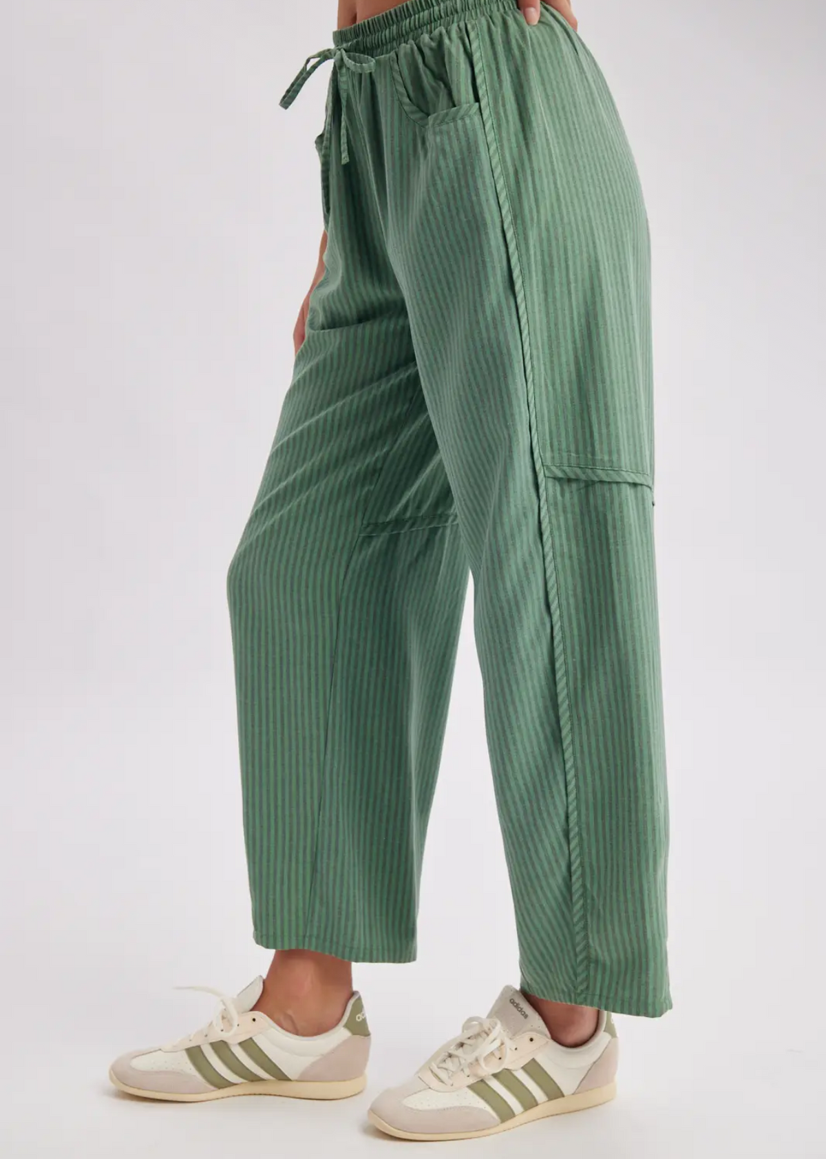 A person wearing sage green pleated wide-leg pants with a drawstring waist and cream sneakers, shown from the waist down against a plain light background.