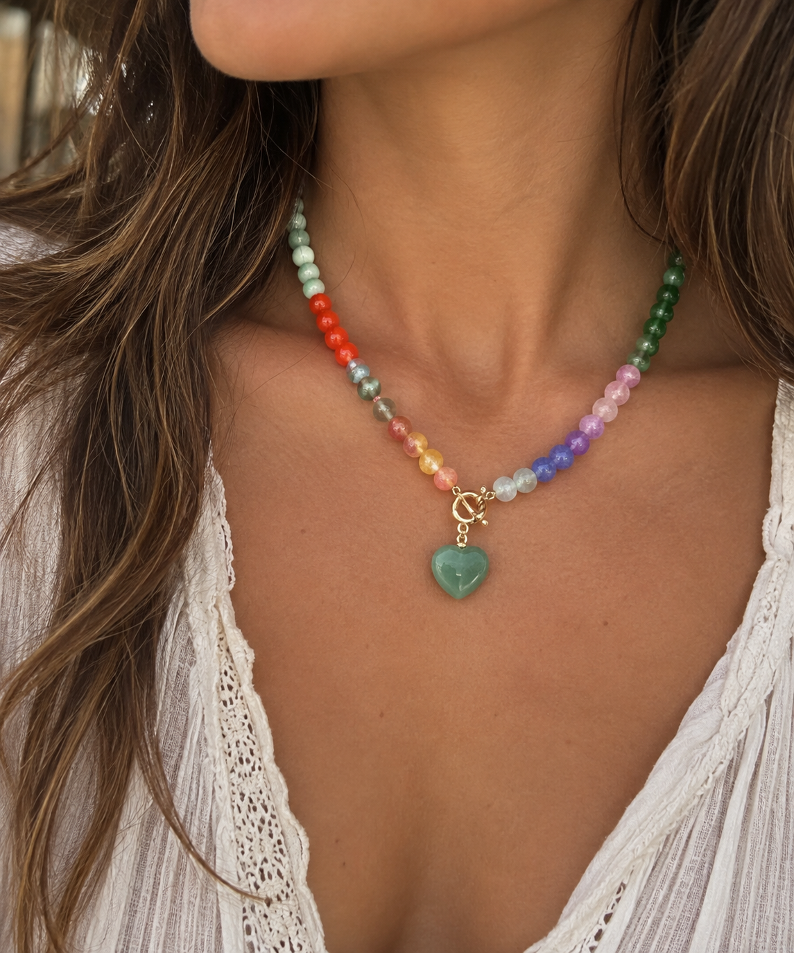 A close-up of a woman wearing a beaded necklace with a green heart charm. The necklace features multicolored beads transitioning through red, orange, yellow, green, blue, purple, and pink, with a gold-tone clasp and crescent moon detail.