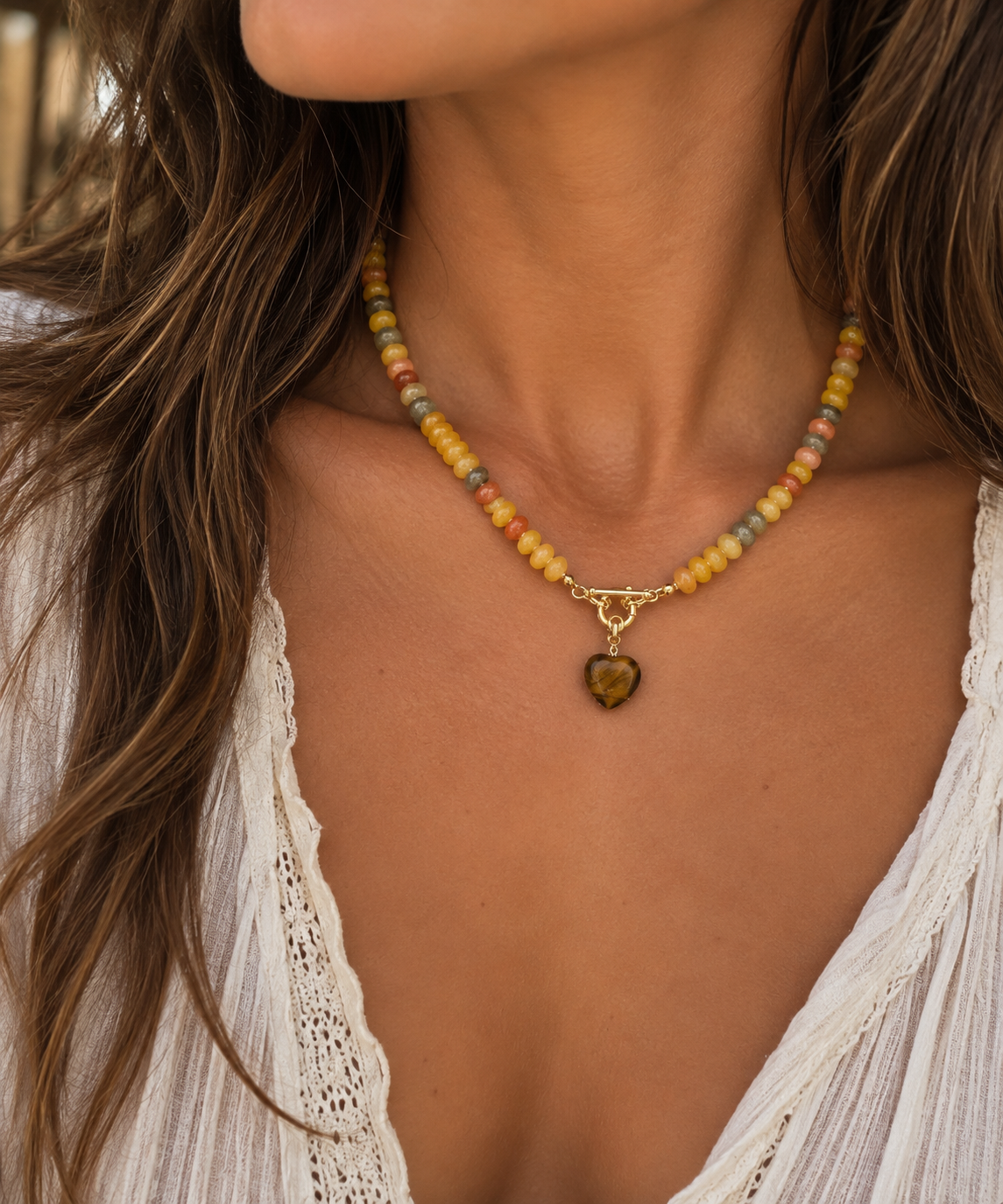 A close-up of a woman wearing a beaded necklace with a gold clasp and a small heart-shaped pendant. The necklace features multicolored beads in shades of yellow, orange, green, and blue, worn over a white lace top.