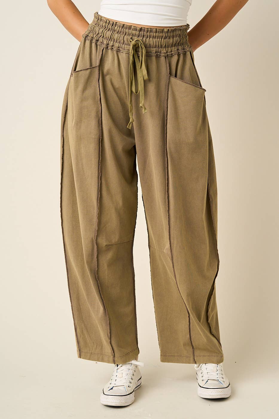 A person wearing loose, wide-leg olive green pants with an elastic drawstring waist, side pockets, and front pleats, paired with white sneakers against a plain beige studio background.