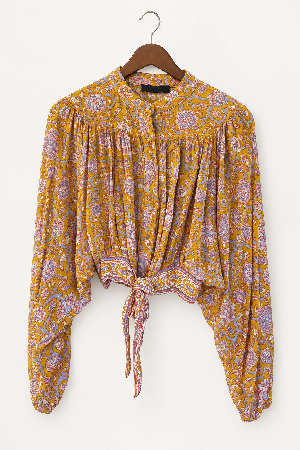 A long-sleeve blouse with a yellow-orange and purple paisley pattern, hanging on a dark wooden hanger against a dark paneled door.