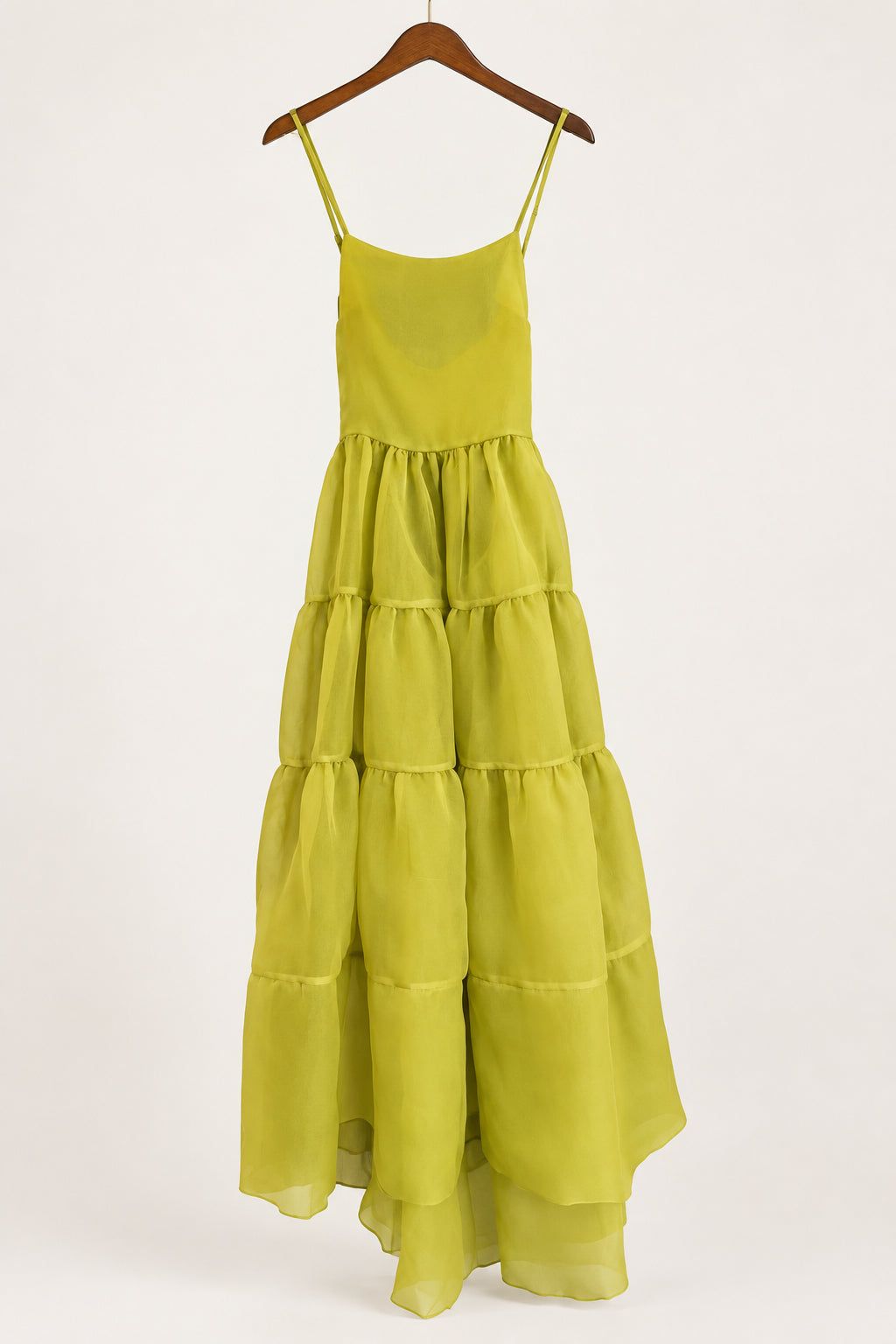 A yellow-green tiered midi dress with thin spaghetti straps, hanging on a dark wooden hanger in front of a dark blue-gray door.