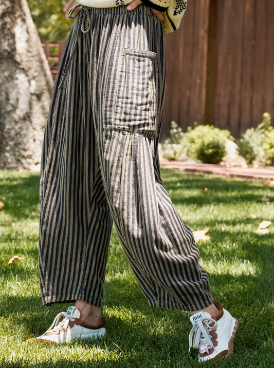 A person outdoors on grass wearing loose, wide-leg striped pants in dark gray and beige, with white sneakers, photographed in a backyard setting.