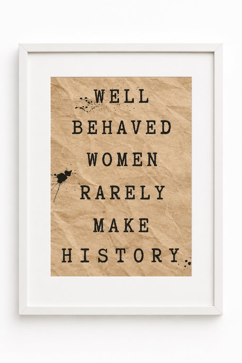 A framed typographic art print with the text 'WELL BEHAVED WOMEN RARELY MAKE HISTORY' on a beige crumpled-paper style background, shown in a clear plastic sleeve on a wooden surface.