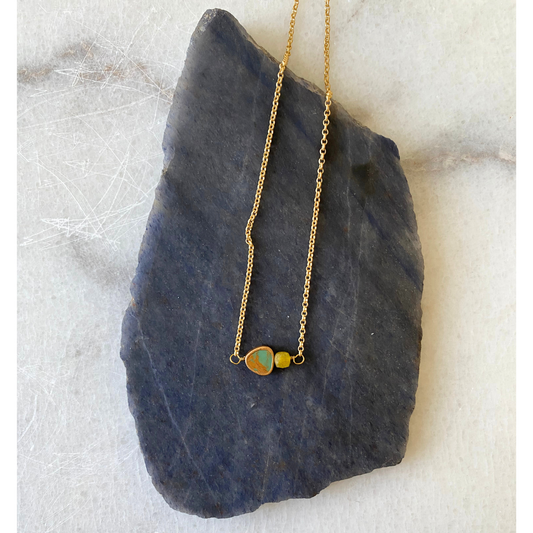 A delicate gold chain necklace with a small pendant featuring a round orange stone and a smaller oval green stone, displayed on a dark slate slab over a light marble surface.