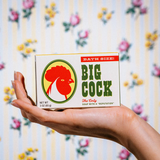 Hand holding Whiskey River Soap Co Big Cock triple milled boxed bar soap against a floral background