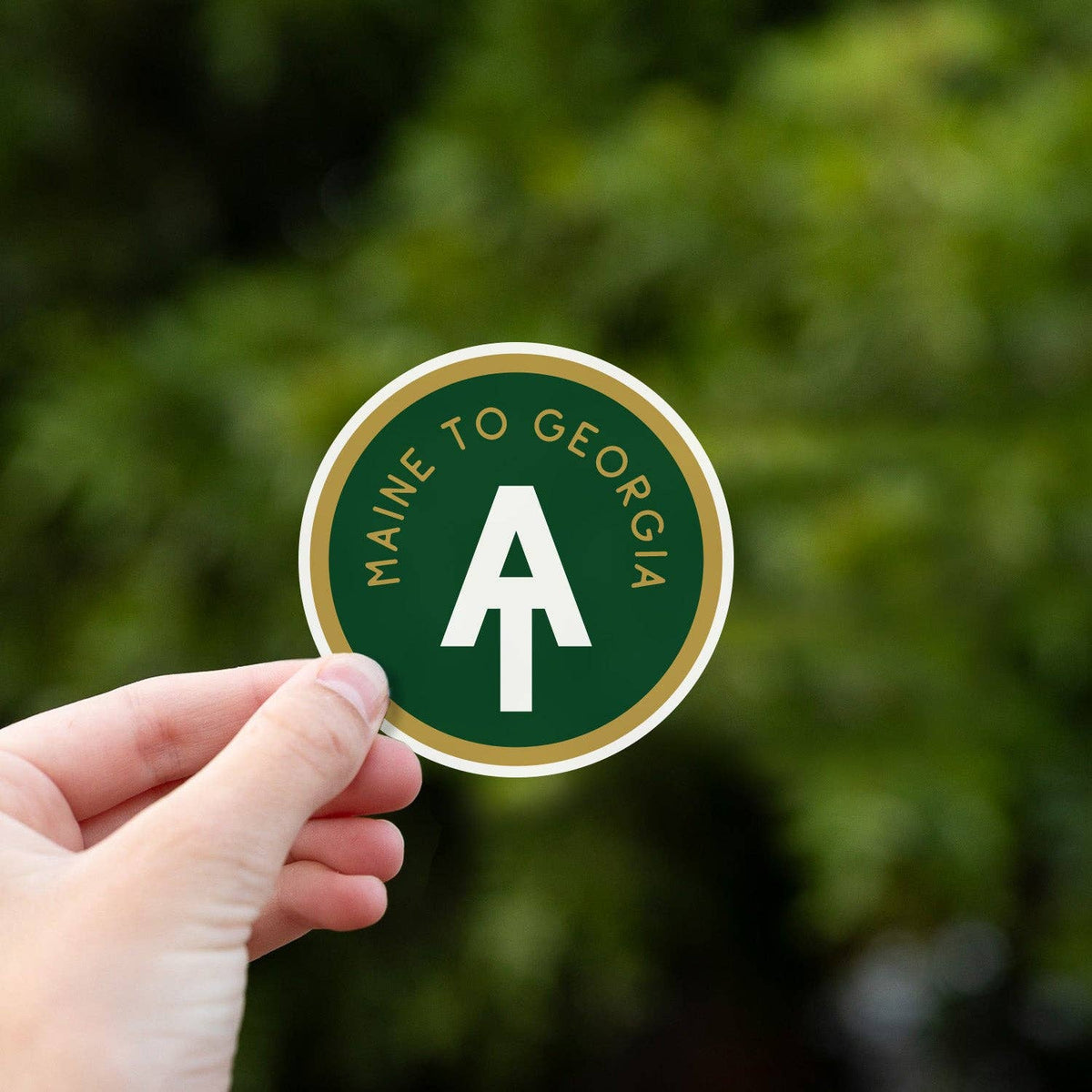 A hand holds a round green sticker with a white arrow and the text 'MAINE TO GEORGIA' against a blurred green outdoor background.