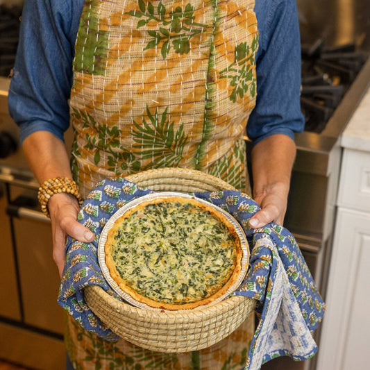 Person holding a pie in a woven basket with a floral cloth, wearing a patterned apron.