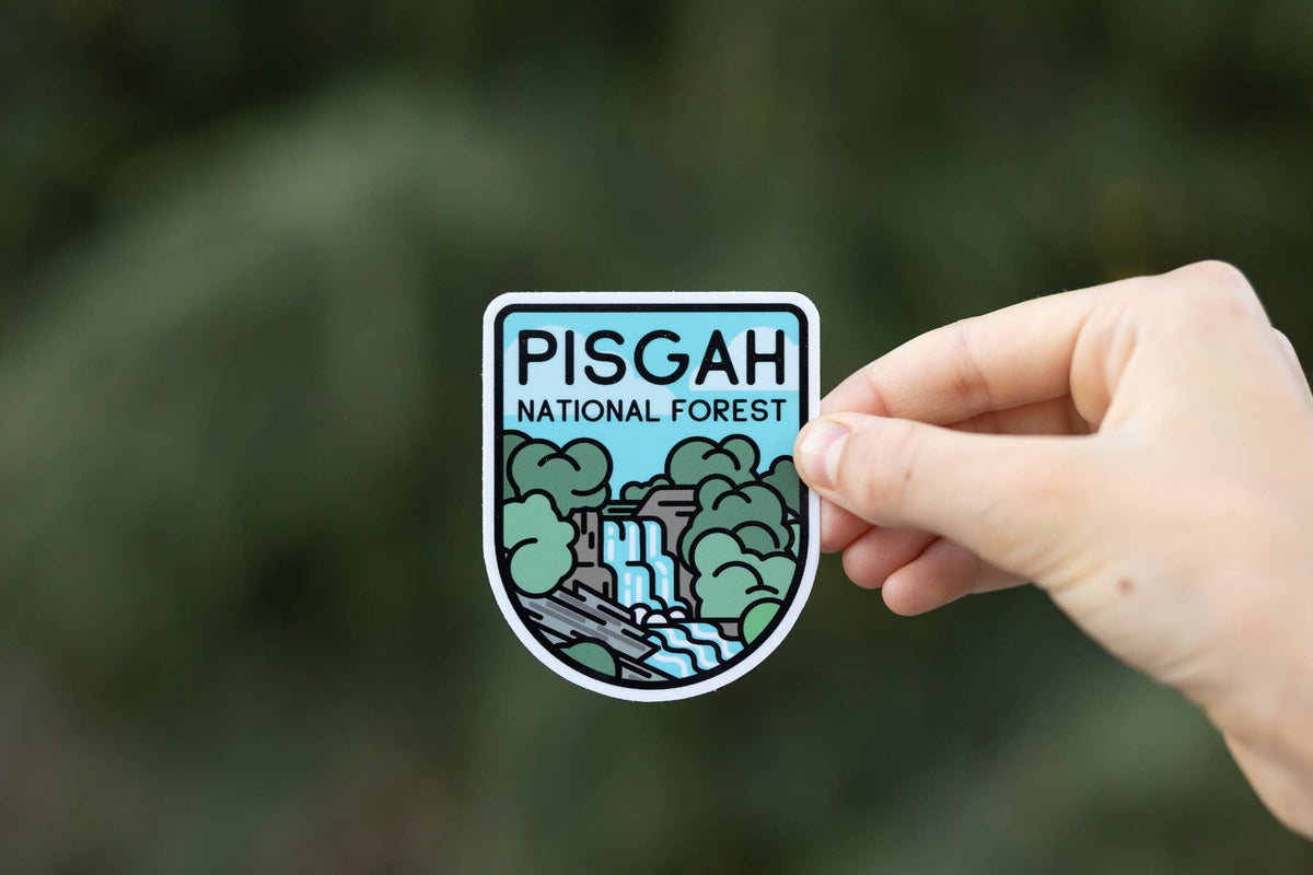 A hand holds a die-cut sticker featuring an illustration of Pisgah National Forest, with green trees, a waterfall, and blue sky, against a blurred outdoor background.